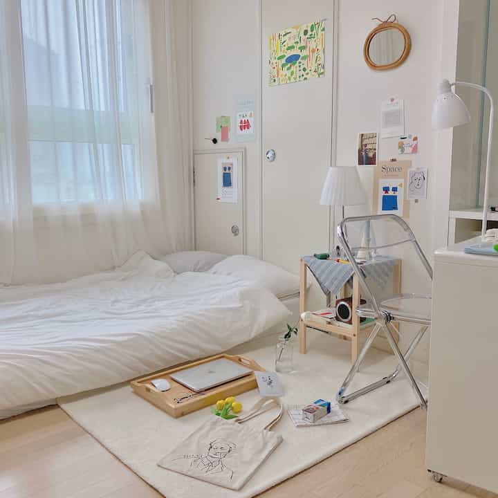 A white and natural color tone studio bedroom featuring a short pile rug, transparent chair, and wooden bed tray with a simple and cozy atmosphere