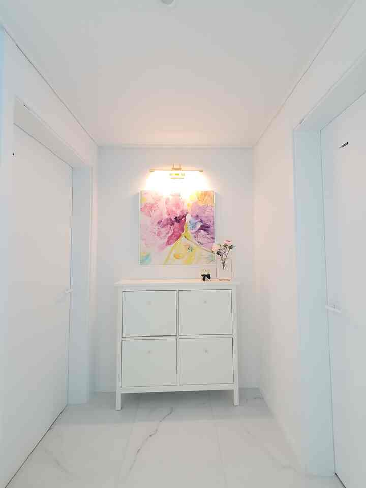 明るいホワイトトーンの玄関、中央に4つの引き出しがあるシューズラックが配置され、上には花瓶とアートが飾られたシンプルなインテリア