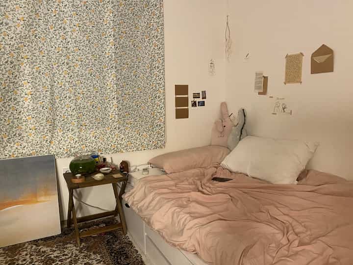 Beige and pink bedding in a studio bedroom with white walls, vintage carpet, and nightstand creating a cozy atmosphere