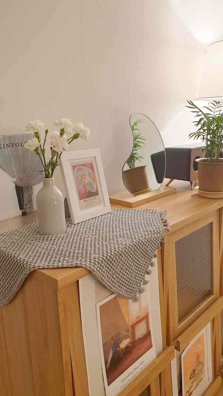 Natural color living room cabinet featuring a vase with flowers, framed art prints, plants, and uniquely shaped mirror in a cozy atmosphere