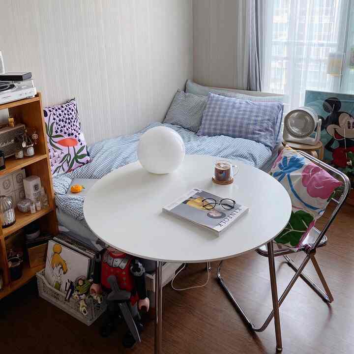 White and brown toned studio bedroom featuring a round table, cozy bed, and neat storage shelves in a compact home cafe setting