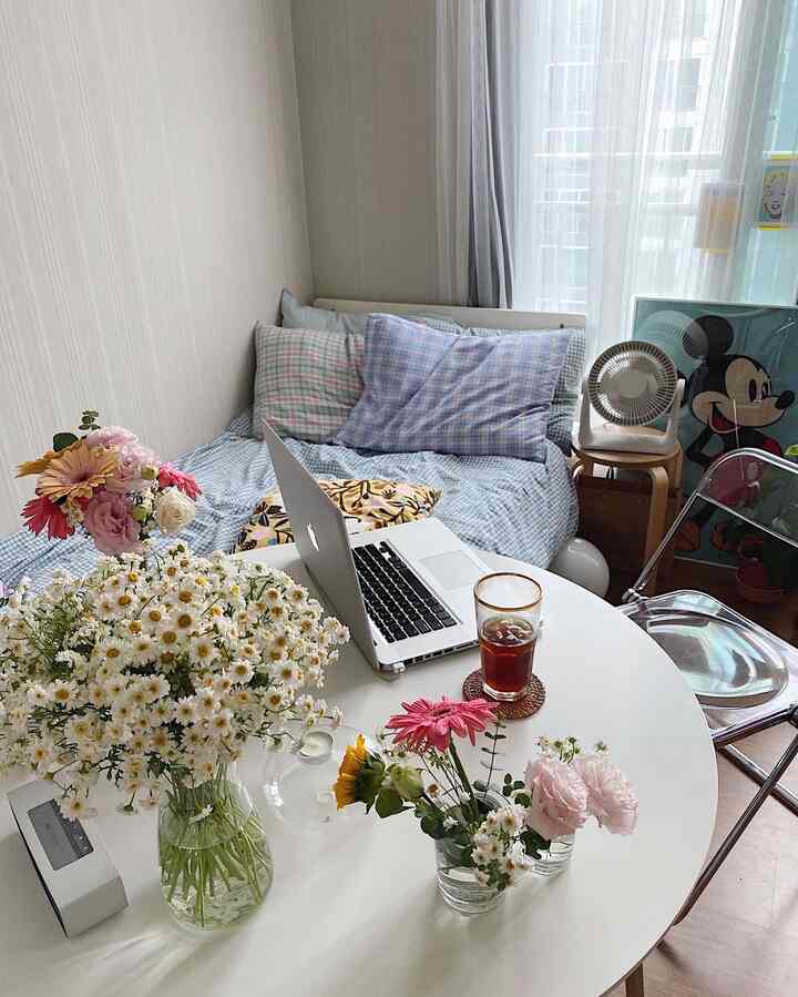 White and natural toned bedroom space with flowers, transparent chair, laptop, and coffee creating a cozy home office atmosphere