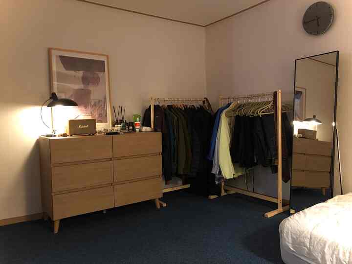 ブルーのカーペットタイル床とナチュラルウッド家具が配置されたワンルーム寝室、洋服ラックと全身鏡のある落ち着いた空間