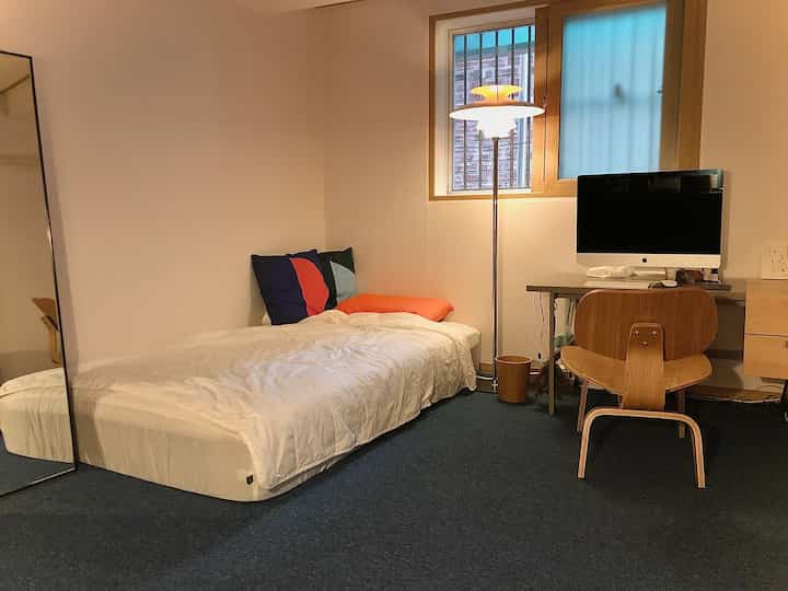 Simple modern bedroom with white walls and blue carpet, featuring a bed, desk with computer, and floor lamp in compact home office setting