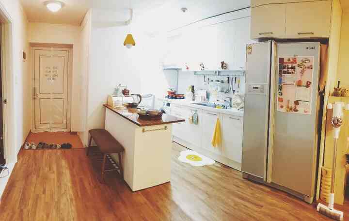 Natural wood-tone floor and white-walled hallway-style apartment entrance and kitchen featuring kitchen island and bench creating cozy space