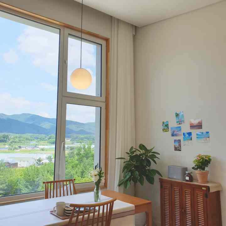 明るいホワイトとウッドトーンの調和が美しい田舎家のリビング、大きな窓とペンダントライト、木製のダイニングテーブルと椅子が配置された雰囲気のある空間