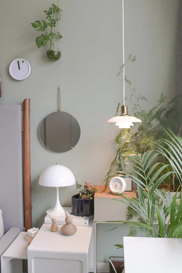 A bedroom with soft green walls and natural materials featuring multiple plants and modern lighting fixtures