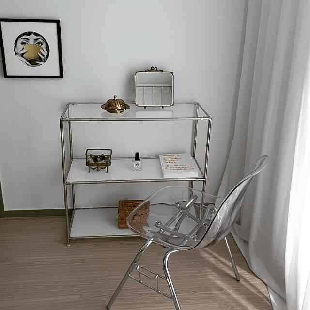 A clean bedroom space with white walls and curtains featuring a metal frame modular shelf and a transparent plastic chair in natural tones