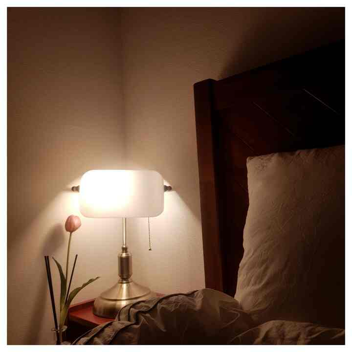 ブラウンのベッドフレームと白い壁が調和したミニマルな寝室、ナイトテーブルの上に置かれたテーブルランプとピンクの花が配された落ち着いた空間