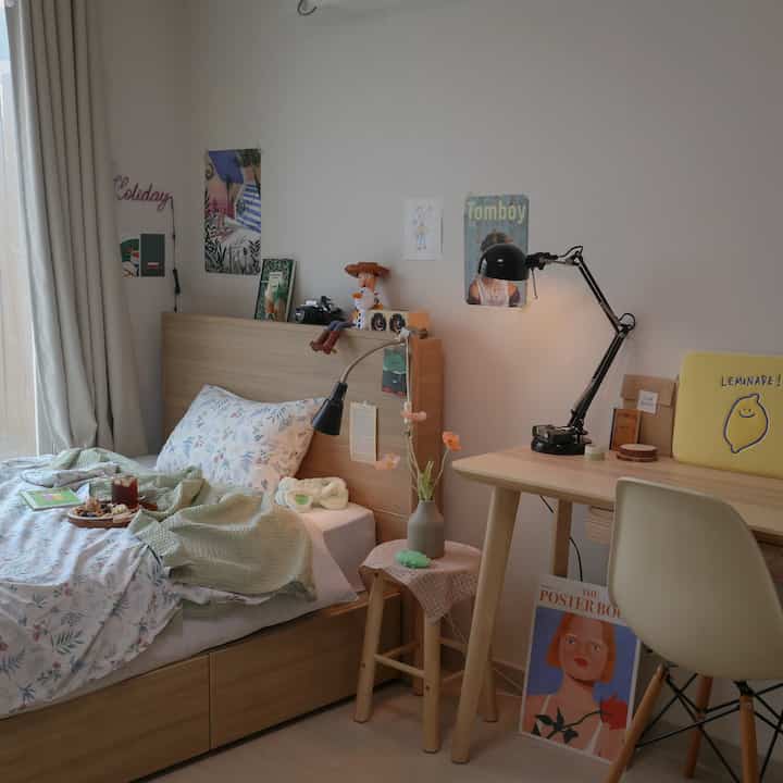 Soft natural wood tone bedroom featuring a wooden bed and desk with table lamp, summer bedding, and posters creating a cozy atmosphere