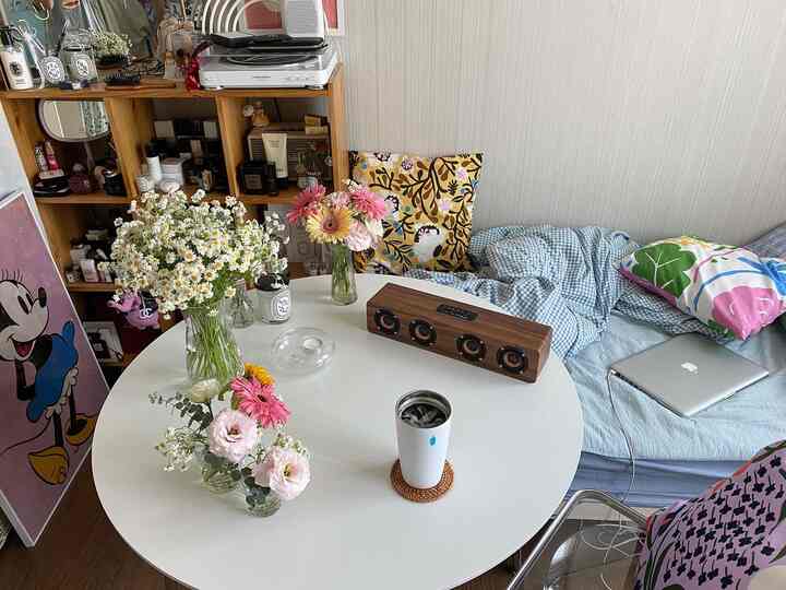 A cozy and stylish studio apartment featuring a white round table, blue-toned bed, vases with flowers, and a Bluetooth speaker for a home cafe vibe