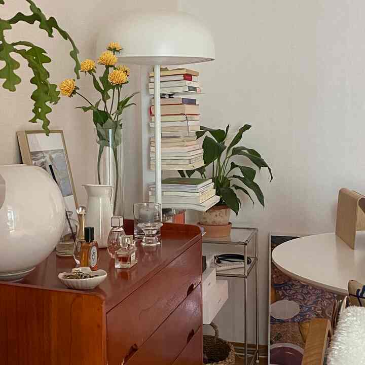 ナチュラルな色調の狭い部屋、木目調の化粧台と白い円形テーブル、フロアランプや植物が配置された落ち着いた空間