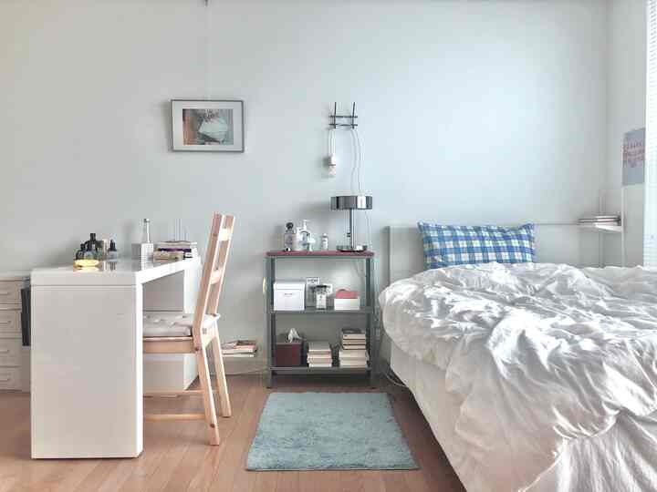 Small bedroom with white walls and natural wood floor, featuring a simple bed and desk, accented with blue checkered pillow and rug for cozy feel
