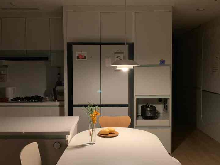 白を基調としたキッチン空間に白いダイニングテーブルとペンダントライトが配置され、黄色の花とマンゴーがアクセントの清潔感あるインテリア