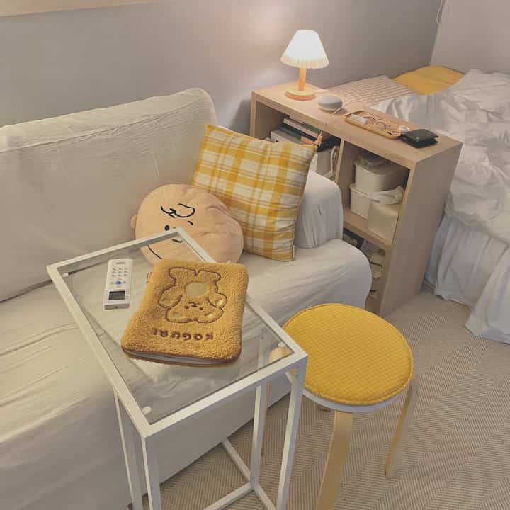 黄色のアクセントを持つ白を基調としたワンルームの寝室、ソファとナイトテーブル、スツールが配置された落ち着いたミニマルな空間