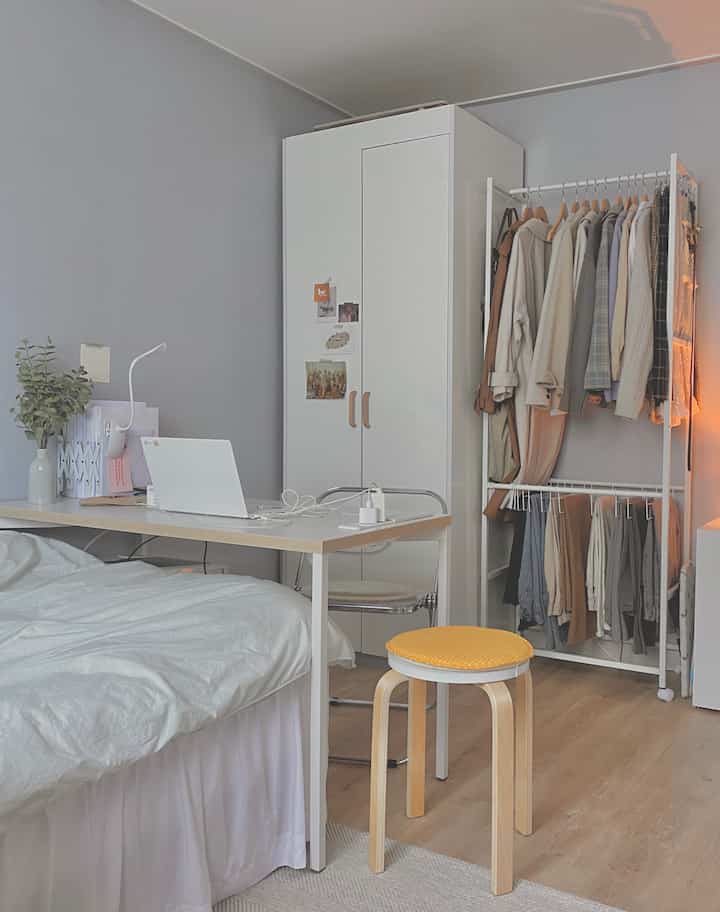 Minimal bedroom in white and wood tones featuring a compact desk with stool, efficient wardrobe and clothing rack arrangement
