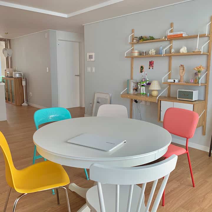 Dining room with light gray walls and wooden shelves, featuring a round white table surrounded by pastel-colored chairs