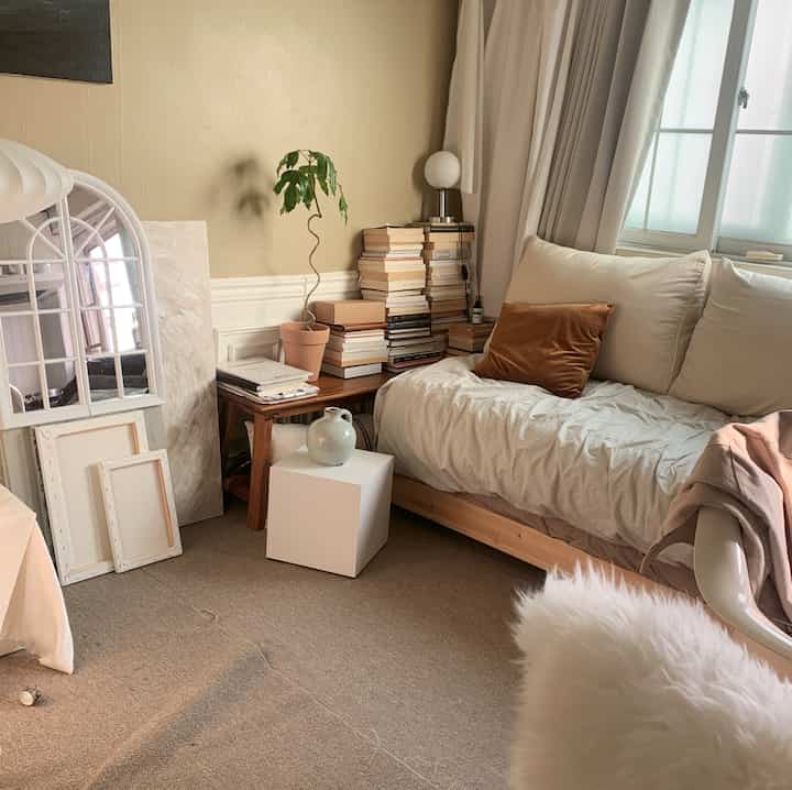 Natural beige and white toned studio bedroom with a nightstand stacked with books and a potted plant creating a cozy atmosphere