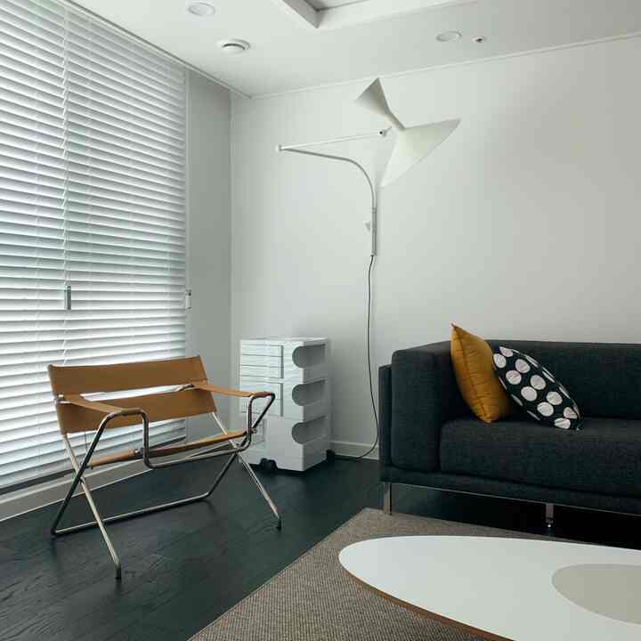 ホワイトとダークグレーのトーンのモダンなリビング、ウッド調のアームチェアとブラインド、シンプルなソファが配置された印象的な空間