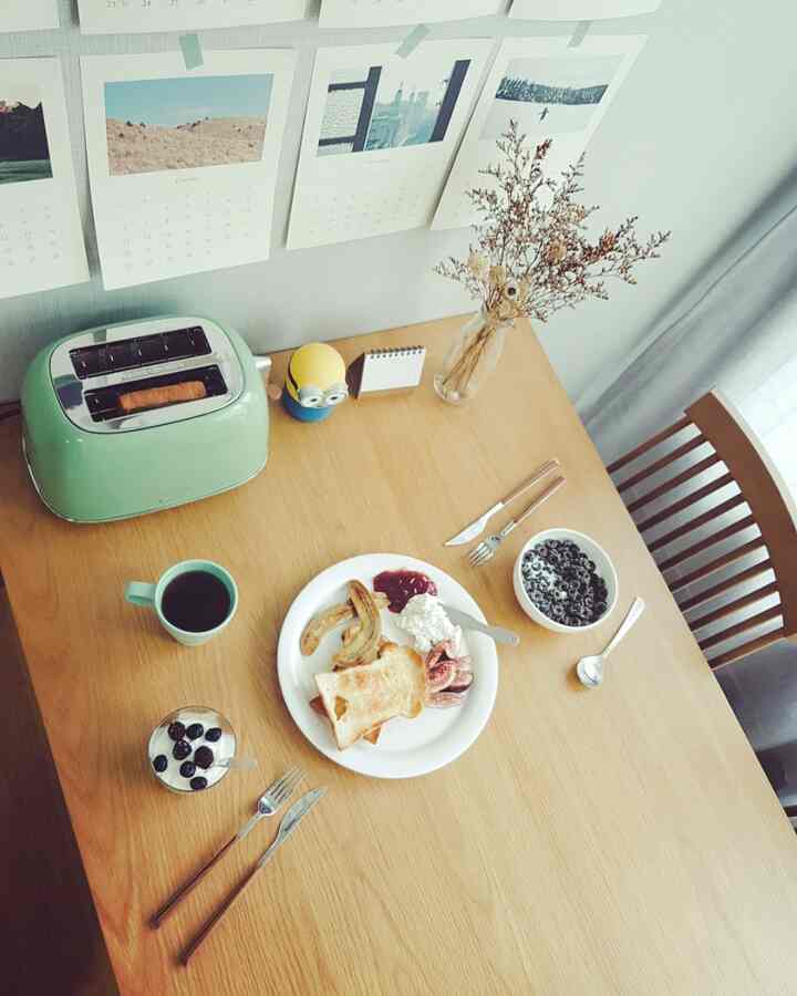 Natural wood-tone dining table near living room window with white curtains, featuring toaster and coffee for a cozy home cafe setting