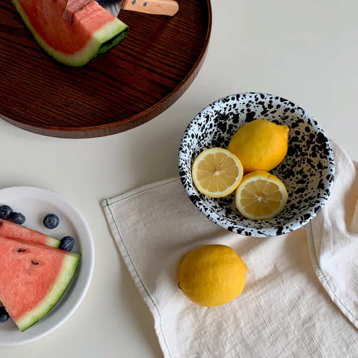 ホワイトのテーブルにウッドトーンのトレイと模様のあるボウルが配置され、スイカとレモンが置かれたナチュラルカラーの清潔なキッチン空間