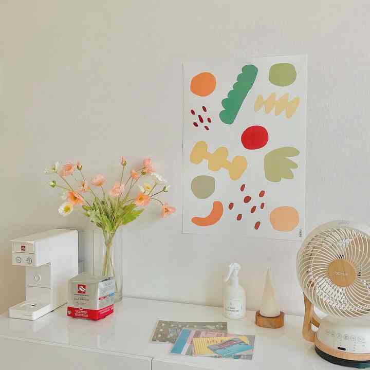 ホワイトトーンのリビングキャビネット上に鮮やかなポスター、ホームカフェ用品、扇風機が配置された雰囲気のある空間