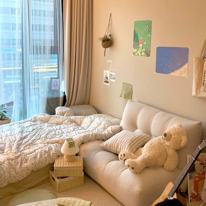 Beige and natural toned 6-pyeong studio living room featuring sofa, bed, curtains, rug, and wall posters with a hanging plant in a cozy atmosphere
