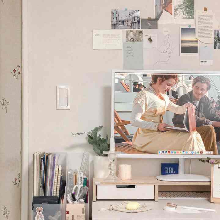 Light wood tone desk and white wall home office space featuring monitor and mood posters, creating cozy remote work environment