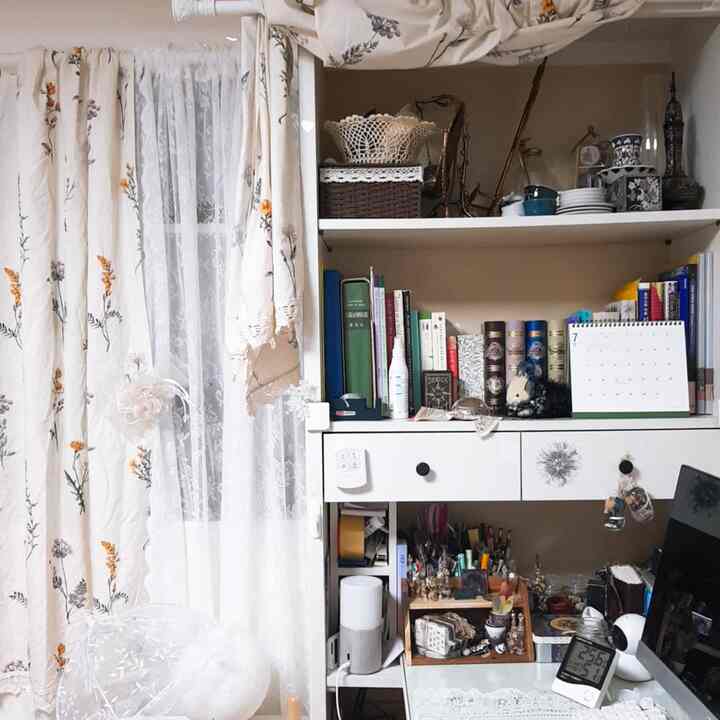 ベージュとホワイトのトーンでまとめられた小さな部屋、花柄カーテンと机、本棚が配置された雰囲気のあるホームオフィス空間