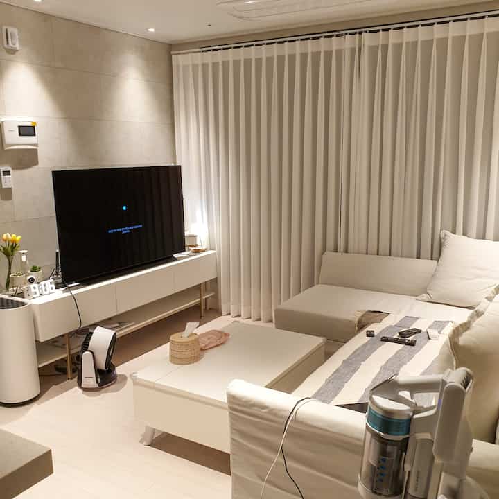 White-toned living room featuring a white sofa, curtains, and TV stand in a clean minimal space