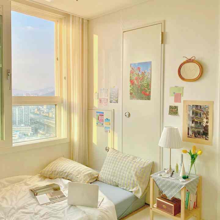 Soft white and yellow toned small bedroom with a large window, yellow tulips, and rattan wall mirror creating a cozy atmosphere