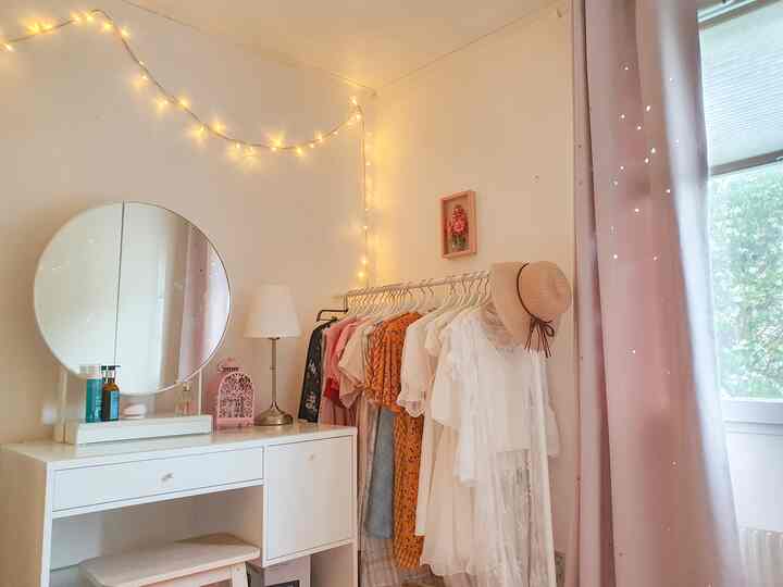 A white and pink toned studio bedroom featuring a vanity, clothing rack, and blackout curtains creating a cozy atmosphere