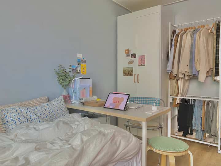 グレーの壁と白い家具が配置されたワンルームの寝室、デスクにタブレットと透明な椅子、クローゼットが調和する居心地の良い空間