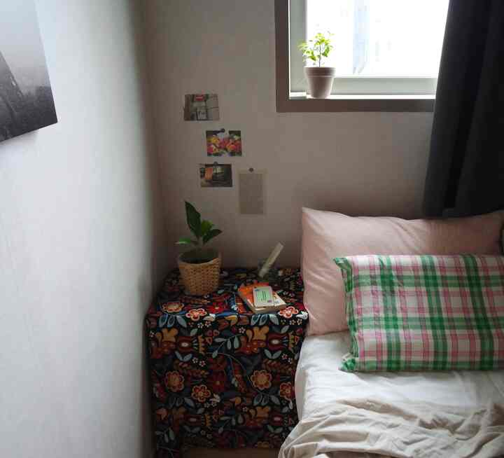 明るいグレーのカーテンと白い寝具、ピンクとチェックの枕が配置された小さな寝室のインテリア