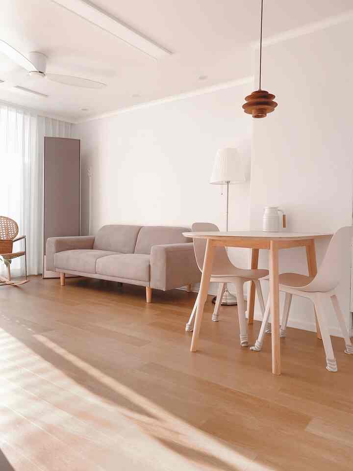 A simple living room with wood-toned flooring and white walls, featuring a gray sofa and natural wood dining table creating a cozy atmosphere