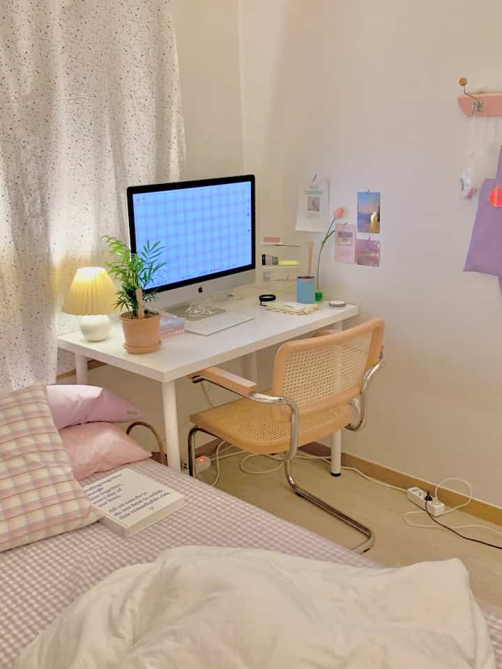 A cozy and tidy bedroom in white and light brown tones featuring a computer desk, bed, plant, and lighting