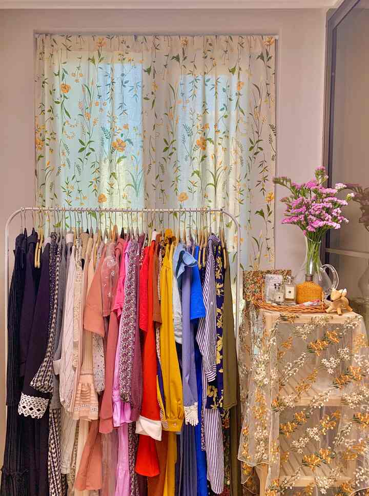 黄色の花柄カーテンと木製棚があるウォークインクローゼット、色別に整理されたワンピースのハンガーとレース布が温かみのある雰囲気を作る空間