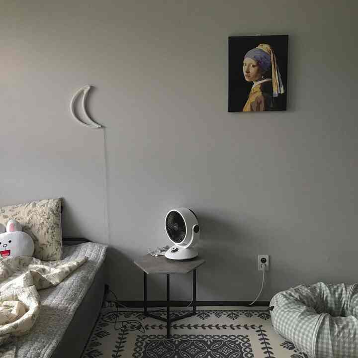 Natural muted-tone small studio apartment featuring a bed, circulator fan on table, and wall art with cozy atmosphere
