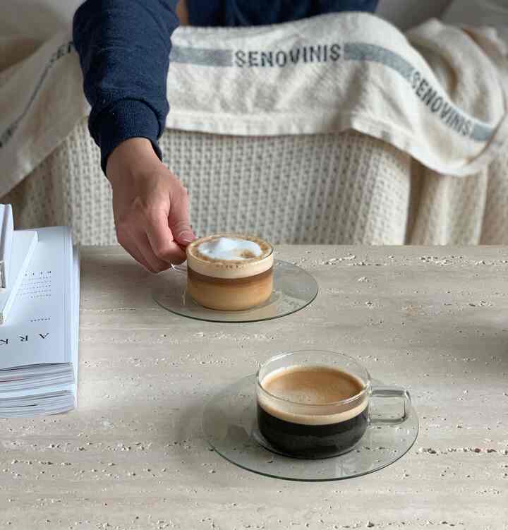 Cozy living room with natural beige tones featuring a stone table with transparent glass plates holding cappuccino and espresso, alongside a Fog Linen throw blanket