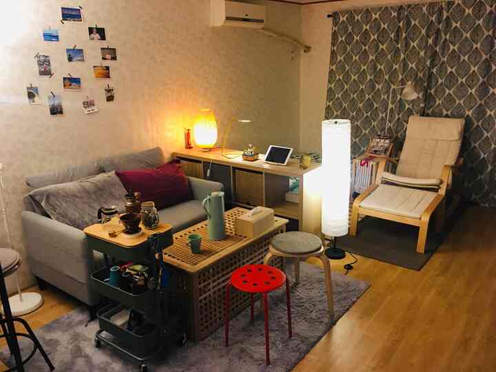 Natural-tone studio apartment living room featuring gray sofa, patterned curtains, multiple lamps, and stools creating a cozy multipurpose space