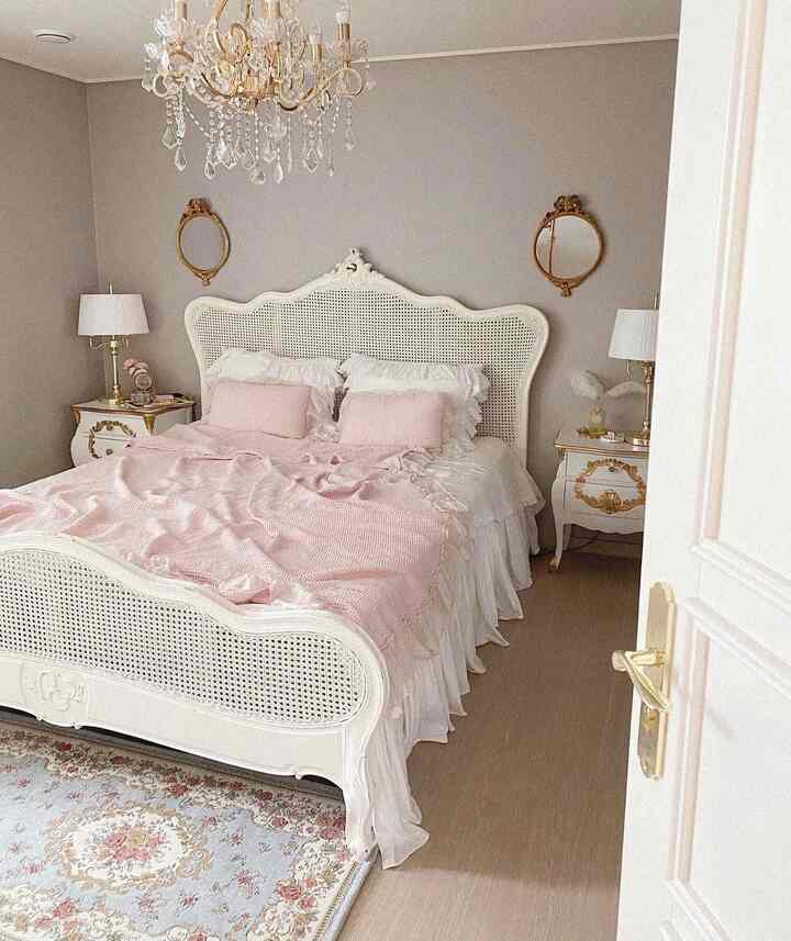 Pink and beige toned classic bedroom featuring an ornate chandelier, antique mirrors, white bedding, and nightstands with elegant decor