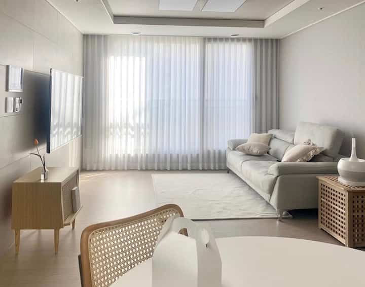 Bright and airy 25-pyeong modern living room in white and wood tones featuring sheer curtains, a sofa, and side table creating a clean ambiance
