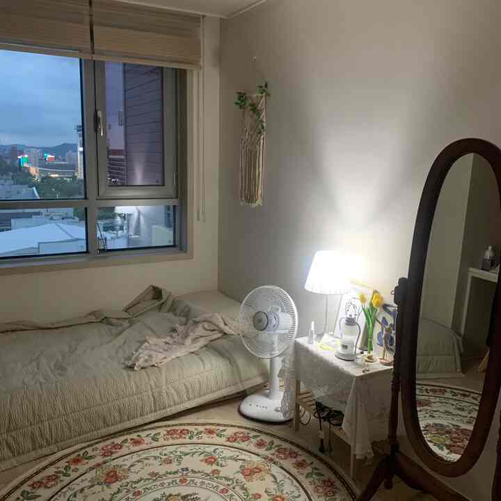 白とベージュの色調の6畳ワンルーム寝室、ベッドと木製の鏡、丸い花柄ラグが配置された雰囲気のある空間