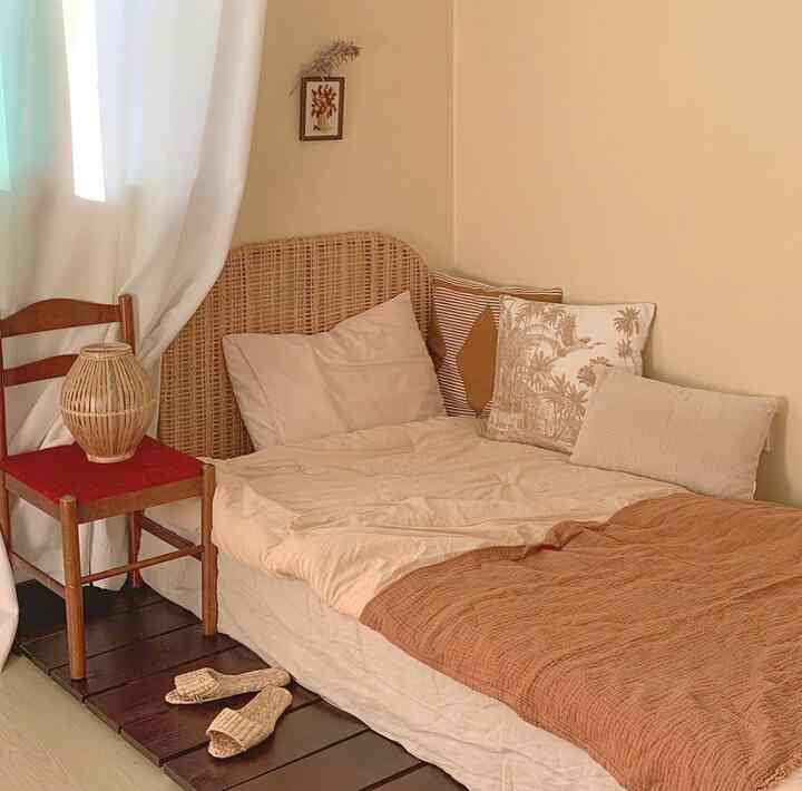 ベージュとブラウンの色調の寝室、ラタンのヘッドボードと多様な枕が配置された雰囲気のあるヴィンテージスタイルの空間