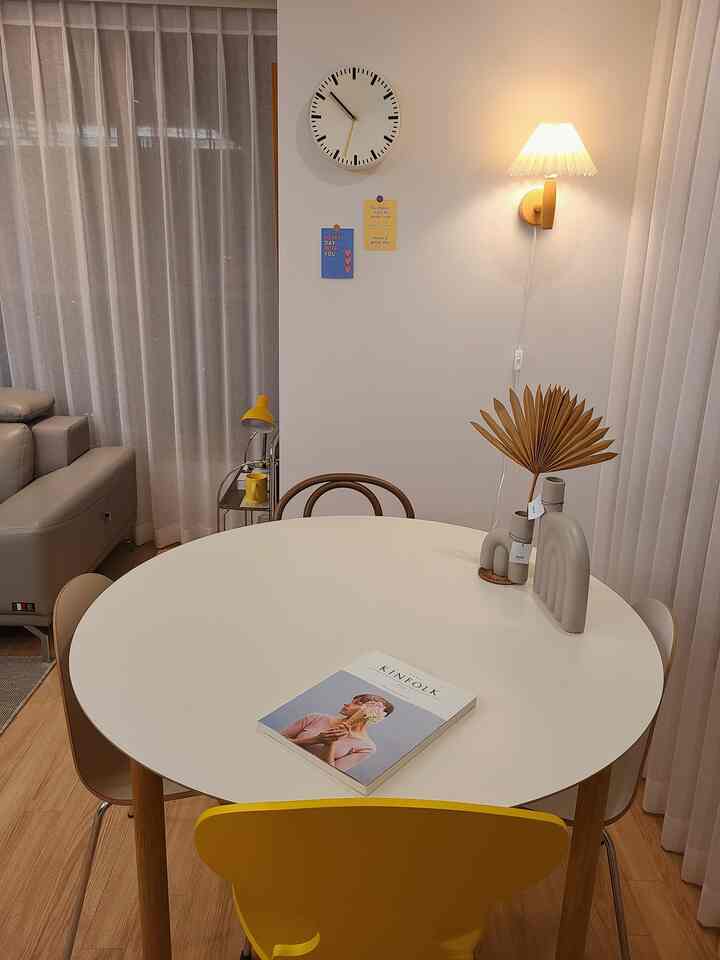 A white and yellow toned dining room featuring a round dining table, curtains, a wall clock, and a wall sconce with a cozy atmosphere