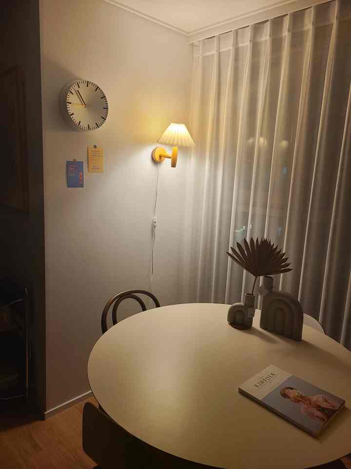 A cozy natural living room with light brown flooring, white curtains, round dining table, and soft wall-mounted lighting