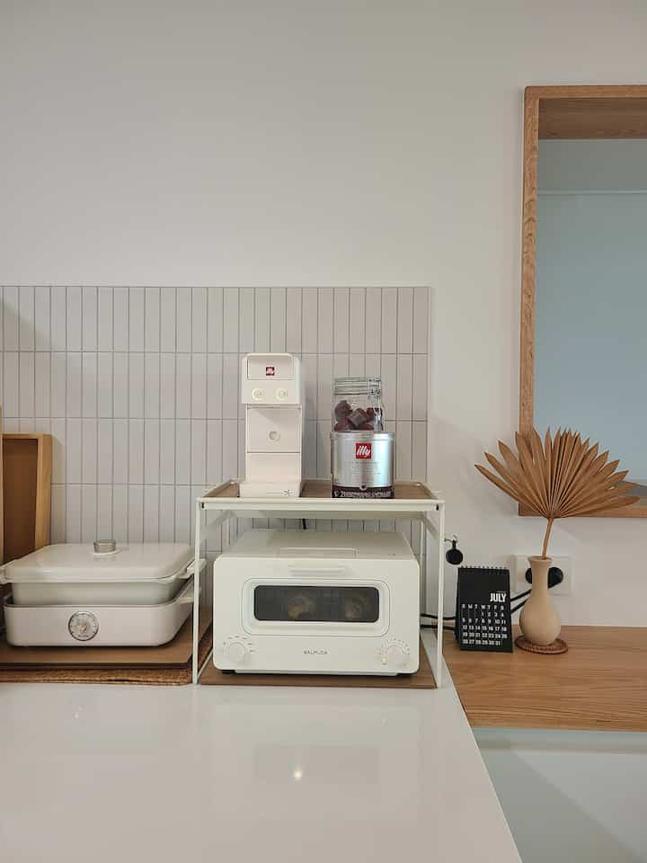 White and wood-tone kitchen featuring a Balmuda toaster and Illy capsule coffee machine in a neat, practical home cafe setup