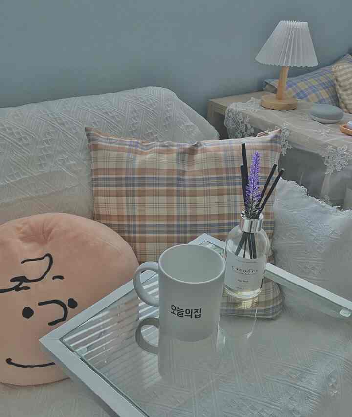 A white and blue-toned living room featuring a white sofa with plaid cushions, glass coffee table with diffuser, creating a cozy atmosphere