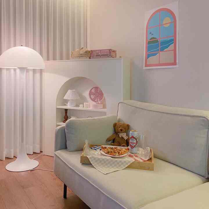 White and natural-toned living room featuring light gray fabric sofa, floor lamp, teddy bear, and tray with casual snack