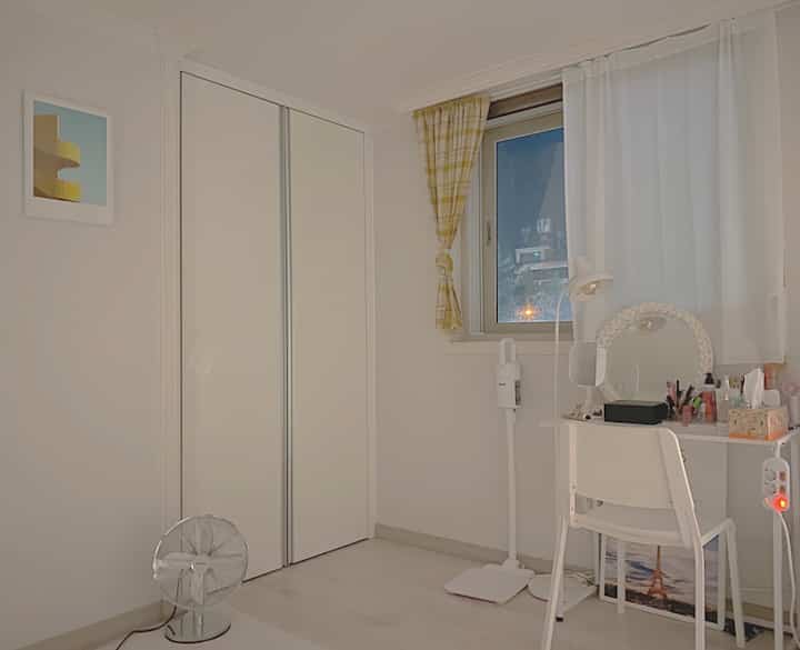 White and yellow toned studio bedroom featuring a neat vanity with floor lamp and checkered curtain, creating a cozy atmosphere